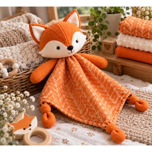May include: A plush fox security blanket featuring an orange and white chevron patterned fabric body with a soft fox head, two arms, and four knotted corners. The fox head has white accents on the ears and face, with black embroidered eyes and a nose.