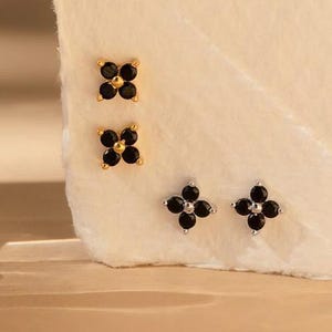 May include: A collection of four pairs of stud earrings. Two pairs feature a gold-tone setting with black gemstone accents in a four-leaf clover design. The other two pairs have a silver-tone setting with black gemstone accents in a four-leaf clover design.