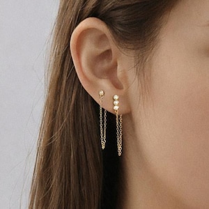 May include: Gold earrings with a delicate design. One earring has a small stud, the other has two studs connected by fine chains. The earrings are worn on the ear of a person.