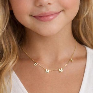 Peut inclure: Une jeune fille blonde aux yeux bleus sourit, portant un collier en or. Le collier comporte les lettres E, M, M et R. La fille porte un haut blanc.