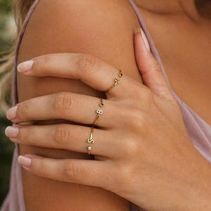 May include: Gold rings with initial letter designs. The rings are worn on the fingers of a person wearing a pink dress. The rings have small gemstones and are delicate in appearance.