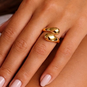 Op de afbeelding: Een goudkleurige ring met een uniek ontwerp. De ring heeft twee druppelvormige elementen die de vinger omsluiten. De ring wordt getoond op een hand met lichtroze nagellak.