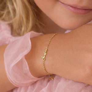 May include: A delicate gold chain bracelet featuring a custom nameplate in a cursive script font. The bracelet consists of a thin, polished gold link chain that connects to the central name element, which is written in a flowing, elegant style.