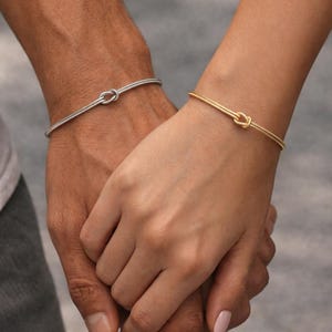 May include: Two hands holding each other, each wearing a thin bracelet with a knot design. One bracelet is silver, the other is gold. The bracelets are simple and elegant, suitable for everyday wear.