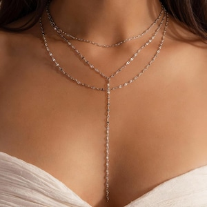 Peut inclure: Un collier argenté à plusieurs rangs avec trois chaînes. Les deux chaînes supérieures sont courtes, tandis que la troisième s'étend sur la poitrine. Le collier est porté sur un haut blanc. Le bijou est délicat et élégant.