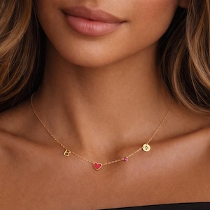 May include: A gold necklace with a fine chain. The necklace includes the letter "B", a red heart-shaped charm, a small pink gem, and a circular charm with a zodiac symbol. The necklace is worn around a person's neck.