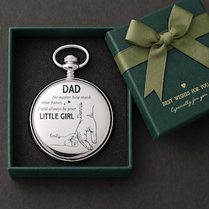 May include: A silver pocket watch with the words "DAD" and "LITTLE GIRL" engraved on the face, along with a drawing of hands. The watch is in a green box next to a gift box with a green ribbon.