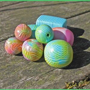 May include: A collection of colourful, patterned clay beads in shades of blue, green, pink, and orange. The beads are arranged on a wooden surface.