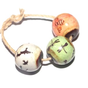 Puede incluir: Una pulsera con tres cuentas de cerámica redondas ensartadas en un cordel de color marrón claro. Las cuentas son blancas con siluetas de pájaros negros, verde claro con motivos de libélulas negras y color melocotón con un diseño de abeja.