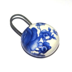 May include: A round button featuring a white base with a blue floral pattern. The button has a dark metal loop attached to the back, extending outward from the circular edge. The surface of the button is smooth and glossy with a distinct rose motif.