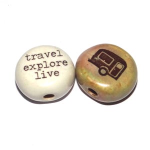 May include: Two ceramic beads with a hole through the center. One bead is white with brown text that reads "travel explore live". The other bead is brown and green with a brown silhouette of a camper van.