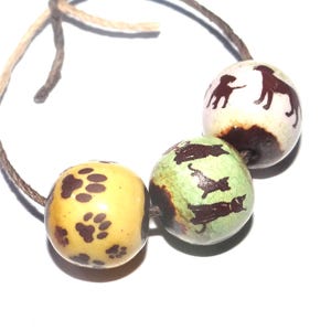 May include: A string of three round beads on a brown cord. The first bead is yellow with brown paw prints, the second is light green with brown cat silhouettes, and the third is white with brown dog silhouettes. Each bead has a dark brown base.