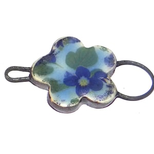 May include: A floral-shaped pendant with a blue and green flower design. The pendant has a metal loop on each side. The flower design is on a light blue background.