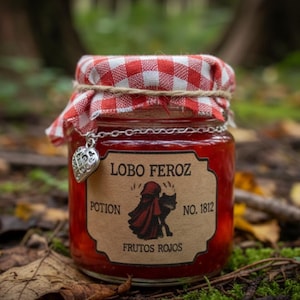 Puede incluir: Un tarro de mermelada roja con tapa a cuadros rojos y blancos, atado con hilo. Un dije de corazón plateado cuelga del tarro. La etiqueta dice "LOBO FEROZ POTION NO. 1812 FRUTOS ROJOS" con un gráfico de Caperucita Roja y un lobo.