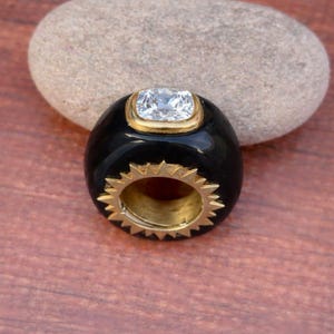 May include: A black ring with a gold-colored interior and a gold-colored bezel around a square, clear gemstone. The ring has a starburst design around the band.
