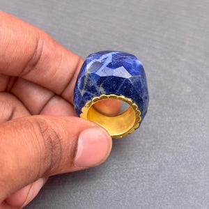 以下が含まれることがあります： ファセットカットが施された深い青色のソーダライトの宝石リングで、研磨された表面に自然な白い筋模様が見られます。石はゴールドトーンの金属バンドにはめ込まれており、リングのセッティングの内周に沿って装飾的なスカラップエッジが施されています。