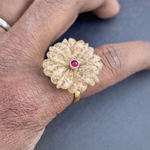 May include: A gold ring featuring a large, carved, beige, flower-shaped stone centerpiece. The center of the floral design is accented with a small, round, vibrant pink gemstone set in a gold bezel, creating a distinct contrast.