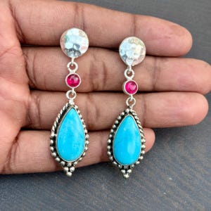 Może przedstawiać: Para srebrnych kolczyków wiszących. Każdy kolczyk ma młotkowany dysk, mały czerwony kamień szlachetny i turkusowy kamień w kształcie łzy z obramowaniem z koralików. Kolczyki są prezentowane na neutralnym tle.