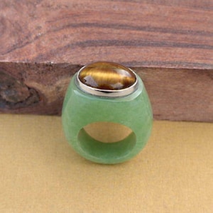 以下が含まれることがあります： 銀色のベゼルにセットされた、大きな楕円形のタイガーアイストーンが付いた緑色の翡翠の指輪。 リングは滑らかで磨かれた仕上げと幅広のバンドを備えています。 タイガーアイストーンは、金茶色と黒のバンドを表示します。