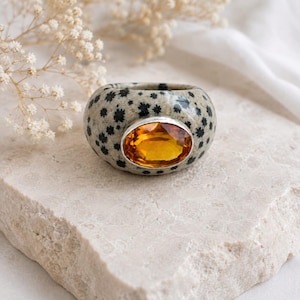 May include: A thick, rounded ring featuring a beige base with a black speckled pattern resembling dalmatian spots. The centre of the ring is set with a large, oval-shaped, translucent amber-coloured gemstone held in a smooth silver-toned metal bezel.