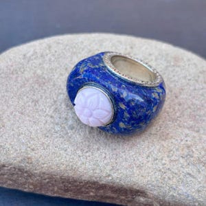 以下が含まれることがあります： 金色の斑点が入った濃い青色のラピスラズリの石が特徴の分厚いリング。リングはシルバーのバンドと、彫刻された薄いピンクの花のアクセントが付いています。リングは灰色の石の表面に置かれています。