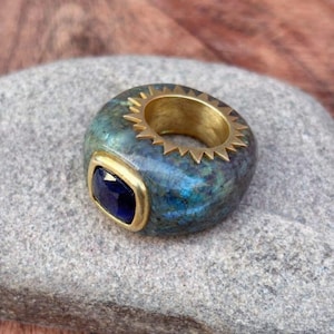 May include: A unique ring featuring a blue-green stone with a gold-colored interior and a gold bezel-set, dark blue gemstone. The ring has a gold sunburst design around the inner edge.