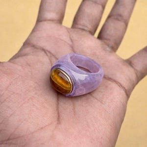以下が含まれることがあります： 薄紫色のバンドと、金色の金属にセットされた大きな琥珀色の楕円形の石が付いた、分厚いリング。 ファッションやジュエリー用のステートメントピースです。