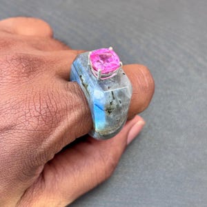 以下が含まれることがあります： 磨き上げられた仕上げと青い虹色の輝きが見えるグレーのラブラドライト石から彫り出されたチャンキーなリング。リングの上面には、4本の爪を持つ銀色の金属製セッティングにセットされた、スクエアカットの鮮やかなピンクの宝石があしらわれています。