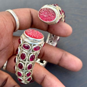 May include: A silver bracelet and ring set featuring carved red gemstones. The bracelet has a cylindrical design with oval-shaped red stones and a floral carved top. The ring has a similar carved red gemstone.