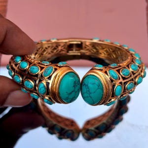 May include: A gold-coloured hinged bangle bracelet adorned with turquoise stones. The bracelet has large turquoise cabochons at the ends and smaller stones along the sides. The design incorporates intricate openwork patterns.