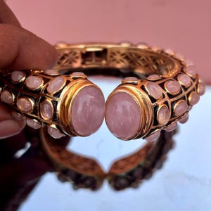 May include: Gold-coloured bracelet with rose quartz stones. The hinged bracelet has large oval rose quartz stones at the opening and smaller stones along the band. The bracelet has a detailed, ornate design with black accents.
