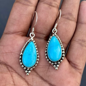 May include: A pair of turquoise teardrop earrings. Each earring has a vibrant blue stone set in a silver-toned frame with a beaded edge. The earrings have a simple hook closure.