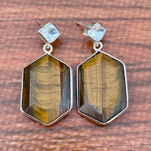 Könnte beinhalten: Ein Paar silberfarbene Ohrstecker. Jeder Ohrring hat einen klaren, quadratisch geschliffenen Stein oben, der mit einem großen, sechseckigen Tigeraugenstein verbunden ist. Die Tigeraugensteine sind braun und goldfarben.