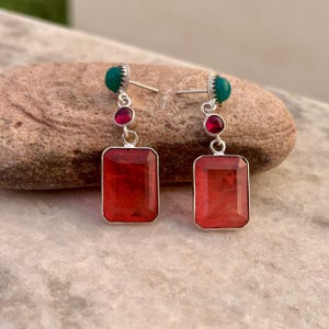 May include: A pair of drop earrings featuring a rectangular red gemstone at the base, a small round red gemstone in the middle, and a small round green gemstone at the top, all set in silver-toned metal with post-style backings for pierced ears.