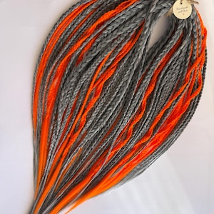 May include: A bundle of grey and orange dreadlocks. The dreadlocks are intricately braided, with alternating strands of grey and vibrant orange. A small tag with the text "Handmade with love" is attached.
