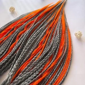 Può includere: Extension per capelli intrecciate grigie e arancioni. Le extension presentano trecce spesse e ben curate in un mix di grigio e arancione brillante. I colori contrastanti creano un effetto visivo audace. Lo stile è moderno e accattivante.
