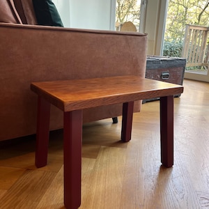 Può includere: Un tavolino in legno con piano rettangolare e quattro gambe. Il piano è di colore marrone caldo, mentre le gambe sono verniciate di rosso scuro. Il tavolo misura circa 46 cm di lunghezza, 30 cm di larghezza e 41 cm di altezza.