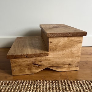 May include: A two-step wooden stool crafted from light brown oak with a visible natural wood grain. The structure features two rectangular platforms at different heights, joined by side panels with visible wooden dowel joinery and a cutout base.