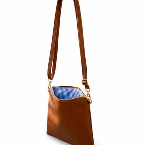 May include: A brown crossbody bag with a long adjustable strap and gold-toned hardware. The bag features a top zipper closure that reveals a blue and white gingham patterned fabric lining inside the main compartment of the rectangular accessory.