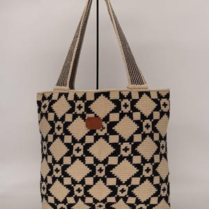 May include: A beige and black tote bag with a geometric pattern. The bag features a repeating diamond and star design. The handles are black and beige striped. A small brown leather patch is centered on the front.