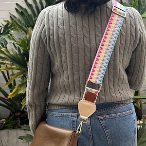 Puede incluir: Una correa de bolso cruzado con un patrón geométrico multicolor vibrante en rosa, amarillo y azul. La correa incluye herrajes de metal dorado, un deslizador ajustable y un punto de unión de cuero beige conectado a un bolso de cuero marrón.