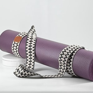 Pode incluir: Tapete de ioga roxo enrolado com uma alça estampada em preto e branco. A alça tem um remendo de couro marrom. O tapete está em uma superfície branca. Um acessório de fitness para ioga e pilates.