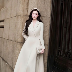 May include: A cream-colored midi-length dress with a notched lapel collar, long sleeves, and a double-breasted button front closure. The dress features a fitted bodice that flares into a full skirt, paired with a small, textured, light-colored handbag.