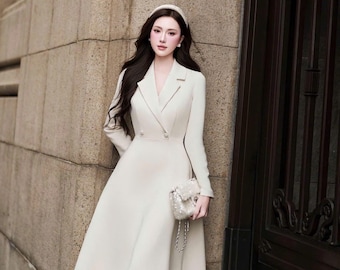 May include: A cream-colored midi-length dress with a notched lapel collar, long sleeves, and a double-breasted button front closure. The dress features a fitted bodice that flares into a full skirt, paired with a small, textured, light-colored handbag.