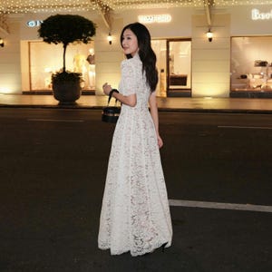 May include: A white lace dress featuring short sleeves and a high neckline. The dress has a full skirt. A person is wearing the dress and holding a black handbag. The background includes shop fronts and street lights.