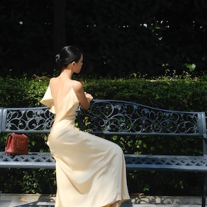 May include: A floor-length pale yellow satin dress with a deep back, thin spaghetti straps, and ruffled shoulder details. The dress is paired with pointed-toe beige high-heeled mules featuring a decorative crystal strap across the top.