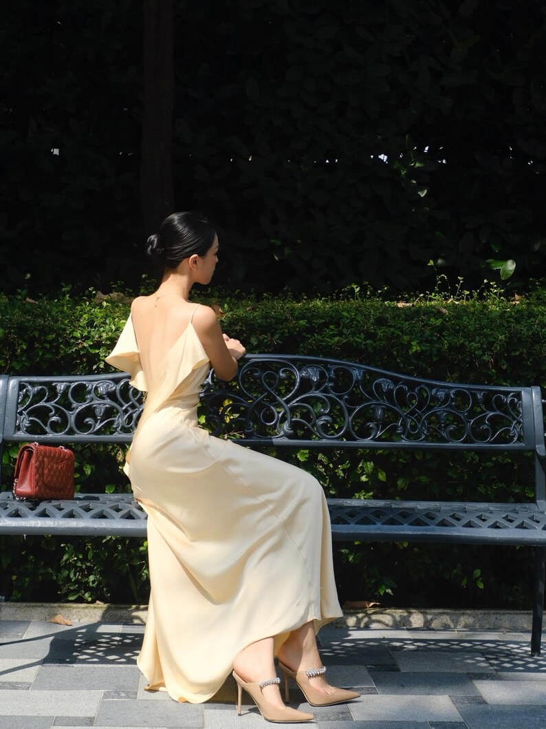 May include: A floor-length pale yellow satin dress with a deep back, thin spaghetti straps, and ruffled shoulder details. The dress is paired with pointed-toe beige high-heeled mules featuring a decorative crystal strap across the top.