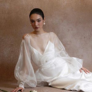 May include: A white wedding dress with a sheer overlay and long, puffy sleeves. The dress features a deep V-neckline and a flowing skirt. A bouquet of white roses lies on the floor. The model is wearing pearl earrings.