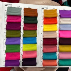 May include: A fabric colour swatch book featuring a grid of small, rectangular textile samples in various colours including yellow, green, pink, purple, blue, red, and brown, organised with numerical labels from 65 to 88 on a white paper background.