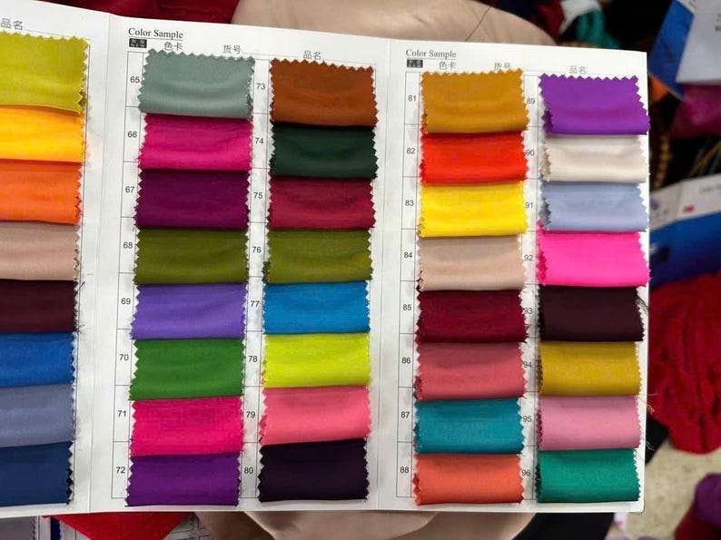 May include: A fabric colour swatch book featuring a grid of small, rectangular textile samples in various colours including yellow, green, pink, purple, blue, red, and brown, organised with numerical labels from 65 to 88 on a white paper background.